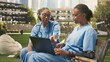© DC Studio - Medical students in blue scrubs discuss information on a laptop while seated on a wooden bench, collaborating with women classmates during outdoor learning and med-school study on campus.