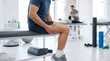 © FajarPutrason - Man holding sore leg while sitting on therapy bench inside bright clinic