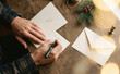 © Nina - Adult man writing a holiday greeting card on a rustic wooden table with envelope and pine branch