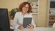 © Krakenimages.com - Woman holding tablet and waving hand at desk in office with monitor and shelves, smiling and wearing glasses while engaging in a remote call; friendly greeting.