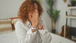 © Krakenimages.com - Middle age hispanic woman with curly hair covering mouth with hands while seated on a bed in a bedroom inside a building; sadness.