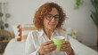 © Krakenimages.com - Woman with curly hair and glasses sipping coffee from a green mug, holding cup with fingers visible while seated on bed in bedroom; calm morning ritual.