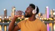 © AZ Studio - A Black man in his 30s wearing headphones drinks water against a blurred city skyline at dusk