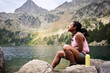 © CarlosBarquero - A Latina African American woman sits on rocks and gazes across a mountain lake with a yellow reusable bottle beside her. Photographed in the Aiguestortes area of the Spanish Pyrenees.