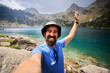 © CarlosBarquero - A Caucasian man smiles and raises an open hand during a lakeside selfie, wearing a hiking hat. Photographed in the Aiguestortes area of the Spanish Pyrenees.