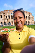 © CarlosBarquero - Vertical. Smiling millennial woman taking a selfie enjoying Italian pizza in front of the Colosseum in Rome, representing authentic travel moments that combine food, culture and happiness vacation.