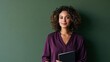 © Prapan - Confident Woman in Purple Blouse Holding a Notebook: A professional woman with curly hair and a warm smile stands confidently against a muted green background, holding a notebook.