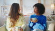 © stockbusters - Multiracial friends drinking coffee sofa. Two women chatting home morning.