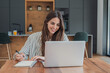 © Daniel - Woman studies online with a laptop and notebook at her kitchen table, writing lesson notes with a pen. Smiling student supports online learning, education, homework, research and study from home.