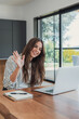 © Daniel - Woman waves to a friend during a long-distance chat on her laptop at home. Her warm smile and hello gesture show friendship, staying in touch, online communication and social connection.