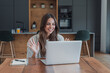 © Daniel - Woman greets colleagues with a wave during a video call on her laptop at the kitchen table. Her smiling home workspace supports remote work, team communication, online meetings and virtual.