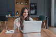 © Daniel - Woman waves to family during a video call on her laptop at the kitchen table. Her warm smile and raised hands portray a virtual family conversation, long distance connection and communication.