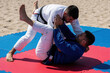 © Vadym - Two men practice jiu jitsu on beach mats. Athletes in blue, white gi fight during outdoor training session. Young men engage in grappling martial arts combat on sand under summer sun. Strong men