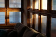 © นิรมล บุญเทพ - cozy indoor photograph capturing warm golden sunlight streaming through a grid of wooden window frames with translucent panels, positioned above a comfortable sofa. This warm composition conveys a ser