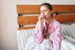 © SHOTPRIME STUDIO - A young woman in pink striped pajamas coughs into her fist while sitting in bed at home. Concept of morning sickness, cold, flu and illness.