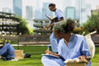 © DC Studio - African American medical student in blue scrubs takes notes while reading a textbook on a campus lawn, preparing for her healthcare studies with books and other academic materials.