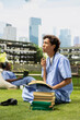 © DC Studio - Thoughtful medical student holds a pen and reviews notes in an open textbook while seated on the campus lawn, surrounded by stacked books and study materials for healthcare education.