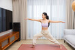 © leungchopan - Woman holding yoga warrior pose on mat in room
