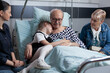 © DC Studio - Cuddling, child and patient rest together at a hospital bedside as the girl leans against a senior man, with family nearby for comfort, affection, companionship, emotional support and recovery.