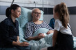 © DC Studio - Conversation, child and patient during a hospital visit as a girl faces a senior man in bed while relatives sit nearby, for communication, companionship, family connection, support and recovery.