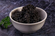© Andrei Starostin - White ceramic bowl filled with glossy black caviar pearls beside fresh parsley on dark textured slate background, luxury seafood delicacy
