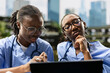 © DC Studio - Medical students, laptop and teamwork frame two classmates reviewing material closely in scrubs. Digital learning, research and peer discussion support collaborative study on an outdoor campus.