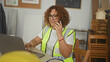 © Krakenimages.com - Woman carpenter talking on smartphone while typing on laptop and reviewing plans, wearing high visibility vest in studio; focused project planning.