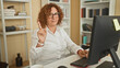 © Krakenimages.com - Curly hair middle age hispanic woman points index finger while typing at desktop computer in office with bookshelves and coffee mug; professional focus.