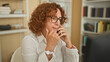 © Krakenimages.com - Woman with curly hair on smartphone with hand at chin and fingers near lips, seated at desk with computer in a building office; quiet concern.