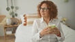 © Krakenimages.com - Woman holding green mug with both hands seated on bedroom bed, smiling and wearing glasses, curly hair visible; contentment.