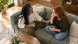 © Christophe - Seen from above, two multiracial friends sit with coffee on a green sofa as one rests a hand on the other’s arm during a quiet conversation in a sunlit living room with a wooden table and plants
