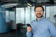 © Tetiana - A close-up photo of a young businessman in a blue shirt and glasses in an office; he is standing, holding a phone, and smiling at the camera