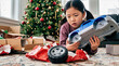 © pufanya - Young East Asian girl looking disappointed while holding a broken blue toy truck with a detached wheel. Christmas morning scene with decorated tree, opened boxes and torn red wrapping paper