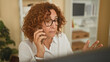 © Krakenimages.com - Woman talking on smartphone, holding device to ear and gesturing with free hand in an office building beside a computer monitor and bookshelf; concern focus.