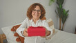 © Krakenimages.com - Woman with curly hair smiling offers red gift box with outstretched hands while seated on bed in bedroom; sharing joy.
