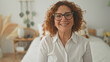 © Krakenimages.com - Woman smiling and adjusting eyeglasses with hands to face while seated on a bed in a studio, wearing white shirt, watch and rings visible; calm contentment.