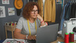 © Krakenimages.com - Woman dressmaker with measuring tape at laptop in studio, hand on chin while working beside sewing machine; quiet focus.
