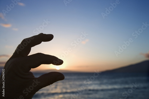 Hand in foreground with fingers around sunrise over coast.