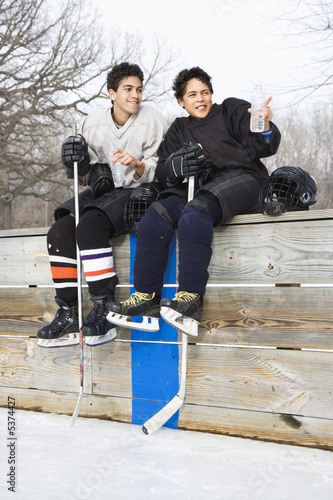 Photography Two boys in ice hockey uniforms sitting on ice rink sidelines.