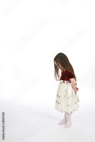 Little girl with head down and hands behind her back.