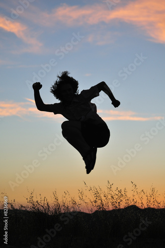 Man jumps high above tall grasses in front of a fiery sunset.