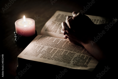 Praying hands on top of bible, lit by candle light