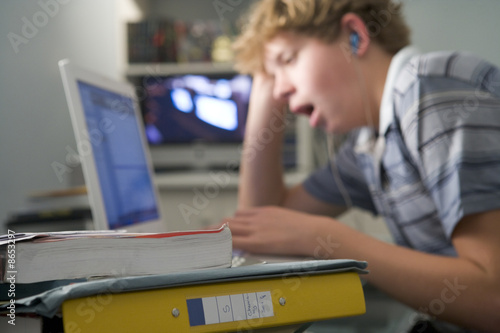 Young boy in bedroom yawning using laptop and listening to MP3 p