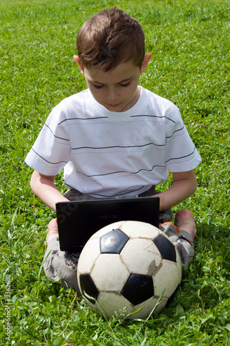 The boy with notebook and a football sits on a grass.