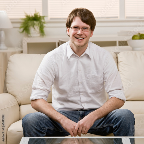 Laughing young man in eyeglasses sitting on sofa at home