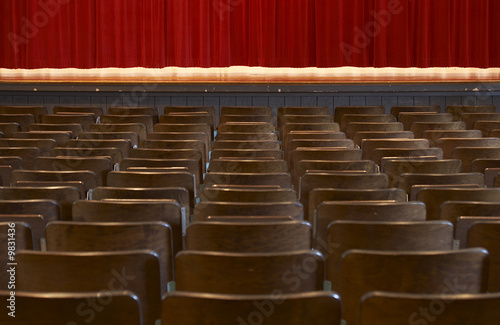 rows of wood chairs in an old auditorium and a red curtain