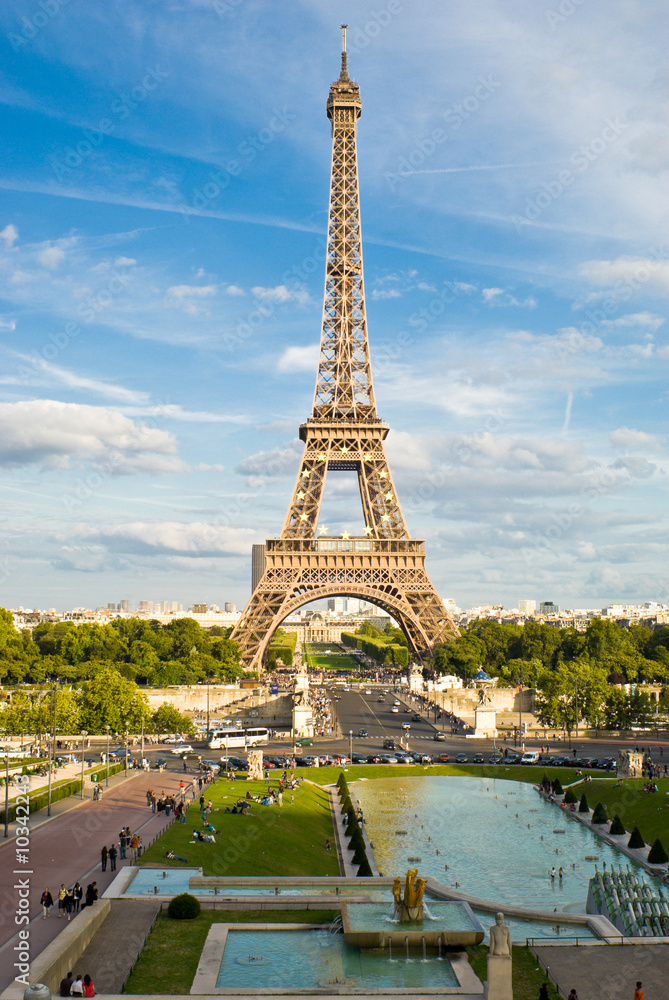 Fototapeta premium Eiffel Tower, with cloudy blue sky and sunny trees around.