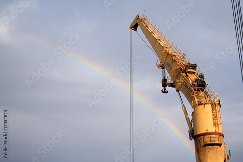 A rainbow on a cloudy sky crosses behind a heavy crane.