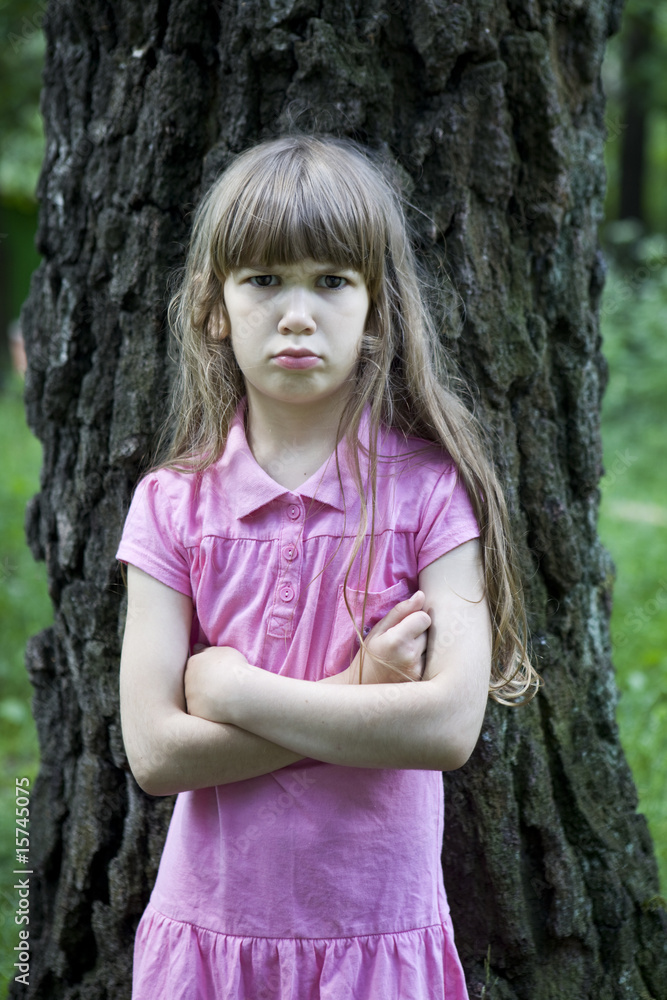 little upset girl wearing pink dress standing near big tree
