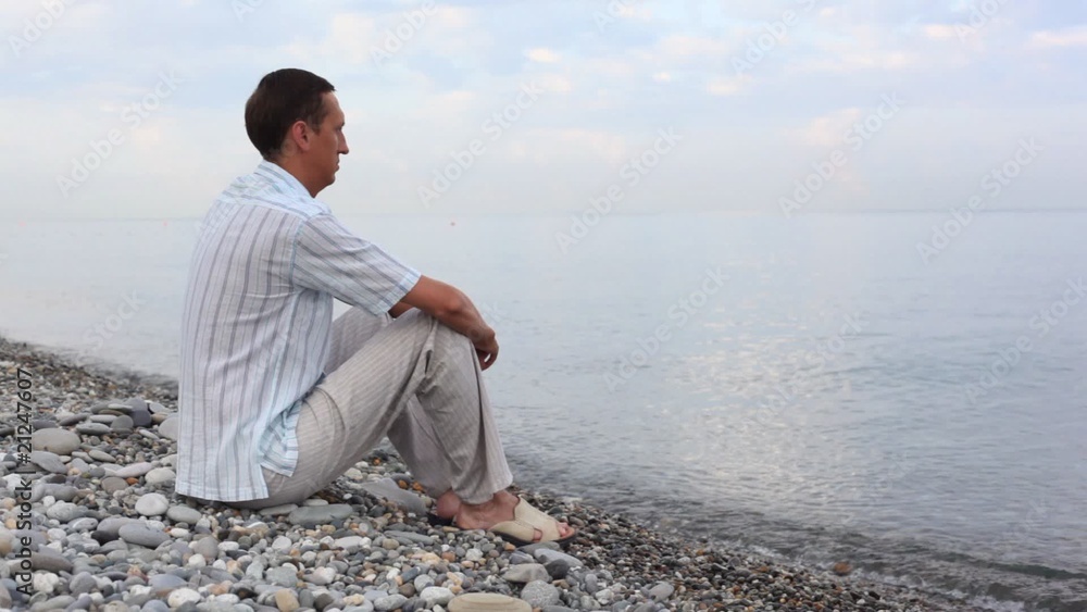 man sitting on pebble coast and looks at sea, side back view man sitting on pebble coast and looks at sea, side back view